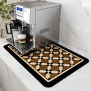 A silver espresso machine with a milk frother sits on a Lovely Home Icon Coffee Machine Mat—a decorative, brown, black, and cream geometric water-absorbent pad with rubber backing—atop a white marble countertop. A cappuccino in a glass cup rests on the drip tray.