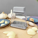 The Lovely Home Icon Manual Pressing Garlic Grinding Grater Cutter Cooking Tool, shown here on a wooden surface surrounded by garlic bulbs, peeled cloves, and two blue patterned plates with garlic, is an essential kitchen gadget for any home chef. Its eco-friendly design combines style and functionality, making it a must-have kitchen accessory from the Kitchen Gadgets and Accessories collection.