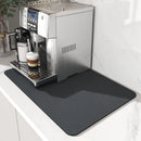 A stainless steel espresso machine brews coffee into a white cup, which sits on the Lovely Home Icon Coffee Machine Mat— a water-absorbent, rubber-backed drying pad—placed on a white countertop against a marble wall, creating a sleek and modern style.