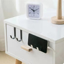 A smartphone charges on a wall-mounted holder attached to a white nightstand, which features black and stainless steel Multi-Purpose Hooks by Lovely Home Icon, a clock, and a wooden lamp on top.
