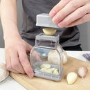 A person uses the Lovely Home Icon Manual Pressing Garlic Grinding Grater Cutter Cooking Tool—an essential kitchen gadget—to mince garlic cloves on a wooden cutting board, with both peeled and unpeeled cloves scattered nearby.