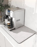 A silver espresso machine brews coffee on a white countertop, with a cup of cappuccino and a mug resting on the drip tray. Enhancing the modern aesthetic, the Lovely Home Icon Coffee Machine Mat—a water-absorbent dish drying pad with rubber backing—adds an elegant touch to the space, while a plant is partially visible in the background.