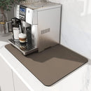 A silver espresso machine rests on the Lovely Home Icon Coffee Machine Mat, a water-absorbent dish drying pad with rubber backing, placed on a white countertop. It brews a beautifully layered coffee in a clear glass mug, creating a tidy and modern look in the kitchen.