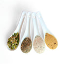 Four white ceramic spoons filled with different Saltopia Low Sodium Salad Seasonings by SALTOPIA Infused Sea Salts are arranged side by side on a white background, showcasing a variety of colorful and textured healthy seasonings.