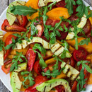 A colorful salad with sliced red and yellow tomatoes, grilled avocado, and arugula on a white plate, finished with SALTOPIA Infused Sea Salts' Saltopia Low Sodium Salad Seasonings for extra flavor.