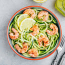 A bowl of zucchini noodles with shrimp and lemon, seasoned with SALTOPIA Infused Sea Salts’ Saltopia Low Sodium Salad Seasonings, sits next to a fork and knife on a light gray surface.