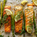 Three baked salmon fillets topped with lemon, fresh dill, and SALTOPIA Infused Sea Salts’ Saltopia Low Sodium Salad Seasonings, arranged on a baking sheet with roasted asparagus and herbs for extra flavor.