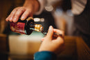 A person pours liquid from a dark glass bottle bearing a red label—likely Taycte Acetaia Malpighi Saba, an Italian cooked grape must syrup and traditional balsamic alternative from the Taycte brand—into a spoon held by another person. The background is softly blurred.