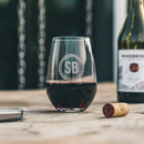 A Stemless Wine Glasses - Set of 4: The Classic engraved with the initials SB inside a circular border, filled with red wine and sitting on a wooden table next to a wine bottle and a cork.