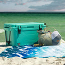 Seafoam 50 quart cooler by Bison Coolers with a mesh bag, a towel, and a hat on the beach.
