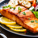 A grilled salmon fillet seasoned with SALTOPIA Infused Sea Salts' PUCKER UP BUSTER Lemon Pepper Salt, garnished with herbs, served with lemon slices, roasted potatoes, and a fresh salad.