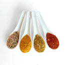 Four white ceramic spoons hold vibrant SALTOPIA Infused Sea Salts—Saltopia Low Sodium Seasonings—in greenish, yellow, light orange, and red on a white background, perfect for low sodium cooking or as a unique seasoning gift set.