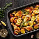A baking dish with roasted chicken drumsticks, red potatoes, lemon wedges, and rosemary sits on a dark wooden table, alongside a bowl of SALTOPIA Infused Sea Salts’ Saltopia Low Sodium Seasonings, garlic cloves, and extra rosemary sprigs.