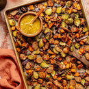 A sheet pan filled with roasted Brussels sprouts, sweet potatoes, sausage slices, red onions, and walnuts is seasoned with Saltopia Low Sodium Seasonings by SALTOPIA Infused Sea Salts. A bowl of mustard sauce and a wooden spoon sit beside an orange napkin.
