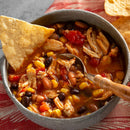 Enjoy a bowl of chicken tortilla soup with shredded chicken, beans, corn, and tomatoes, topped with a tortilla chip. Made with Saltopia Low Sodium Seasonings by SALTOPIA Infused Sea Salts for flavorful, low sodium cooking. A spoon rests on a red striped cloth.