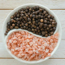 A white round dish in a yin-yang design holds black peppercorns and Pink Himalayan salt from the Saltopia SALT SNOBS Gift Set by SALTOPIA Infused Sea Salts, displayed elegantly on a light wooden surface.