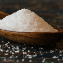 A wooden spoon holds a heap of SALTOPIA Infused Sea Salts from the Saltopia SALT SNOBS Gift Set, with some salt crystals scattered on a dark surface below.