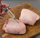 A hand uses the SpitJack SQUIRT 50 Meat Injector Kit with Case and Two Needles to inject marinade into raw chicken thighs on a wooden cutting board.