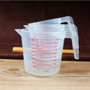 A stack of three Lovely Home Icon plastic measuring volume beakers (250ml, 500ml, and 1000ml) with red PVC metric markings rests on a wooden surface; a wooden stick is partially visible behind them against a brown background.