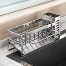 A Lovely Home Icon Sink Cleaning Tools Storage Rack sits on the edge of a kitchen sink next to a window. This multifunctional stainless steel drain rack features an open-grid design for drying dishes and utensils, and includes built-in holders for sponges, cleansers, and rags to keep your kitchen organized and tidy.