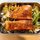 Two pieces of salmon, topped with SALTOPIA Maple Garlic & Thyme Salt: MAPLE ME CRAZY, rest on asparagus and sliced mushrooms in a metal roasting pan, ready for cooking.