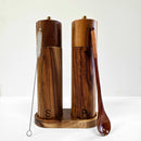 A pair of Spice Wheel Acacia Wood Salt and Pepper Grinders, distinguished with S and P labels, are displayed on a matching acacia wood tray. The adjustable coarseness of the pepper grinder ensures precise seasoning. A brush is propped against the left grinder, while a wooden spoon rests beside the right one—all showcased against a clean white backdrop.