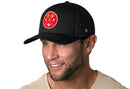 A man smiles wearing a black Haka Hat San Francisco Trucker Hat, featuring a circular red and gold patch with a bridge, "SF," and a football.