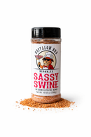 A plastic shaker bottle of BuffalowBBQ Sassy Swine BBQ Rub featuring a label with a cartoon pig in a chef’s hat and sunglasses, sitting in a small pile of the seasoning.