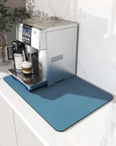 A silver espresso machine rests on the Lovely Home Icon Coffee Machine Mat—a blue, water-absorbent dish drying pad with a rubber backing—placed on a white countertop. A cup of coffee sits beneath the spout, with a plant in the background, creating an inviting and modern table decoration scene.
