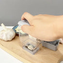 A hand presses down on the Lovely Home Icon Manual Pressing Garlic Grinding Grater Cutter Cooking Tool, filled with garlic cloves, atop a wooden board. Nearby sit whole garlic bulbs, a blue dish with more cloves, and stainless steel blades—making this eco-friendly kitchen gadget both practical and sustainable.