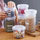 Six Lovely Home Icon Plastic Sealed Tank Large Capacity Crisper Kitchen Organize Grain Storage Boxes with Scales display a variety of dried foods—beans, seeds, nuts, and fruit—in their clear jars. On a wooden surface, a person’s hands are placing an airtight Spice Container holding dried fruit on top of other stacked sealed boxes, demonstrating the practical use and organization these transparent containers offer in the kitchen.