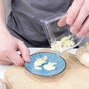 A person pours thinly sliced garlic from the Lovely Home Icon Manual Pressing Garlic Grinding Grater Cutter Cooking Tool onto a blue patterned plate, which sits on a wooden cutting board, demonstrating this eco-friendly kitchen gadget and accessory in use.