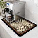 A silver espresso machine sits on a white counter, with the Lovely Home Icon Coffee Machine Mat—a water-absorbent, rubber-backed dish drying pad in a black-and-beige pattern—placed underneath. A glass of foamy cappuccino rests on the machine’s drip tray, while a plant in the background adds a touch of modern decor to the kitchen scene.