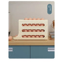 A cream-colored Lovely Home Icon Refrigerator Egg Storage Box with four layers of automatic rolling egg holders sits on a kitchen countertop above blue cabinets, storing rows of brown eggs. This practical fridge organizer dispenses eggs conveniently from the bottom row.