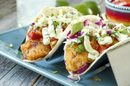 Two soft tacos with grilled fish, cabbage, tomatoes, avocado, and cilantro, topped with Terrapin Ridge Hot Wasabi Garnishing Squeeze by Terrapin Ridge Farms. Served on a blue plate with lime wedges in the background.