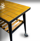 A kitchen cart with a glossy, mustard-yellow tiled top and a black metal frame—echoing the modern style of the WPPO LLC Direct Infinity Side Table Long Side 103cm, designed to fit Infinity 50 and 66. It features a slatted wooden lower shelf and stands on black metal legs with an exposed caster wheel for easy mobility, all set against a white background.