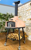The WPPO LLC Direct WPPO Wood Fired, Stone Oven for Bread not Pizza! Red Rock 100 features a tall chimney and rests securely on black wheeled legs. Firewood is neatly stacked below this versatile bread oven, which is placed on a tiled patio framed by a stone wall and vibrant green plants in the background.