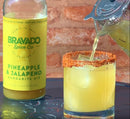 A glass rimmed with chili powder is filled with a vibrant yellow drink poured from a shaker. Beside it sits a bottle of Bravado Spice Co Pineapple & Jalapeño Margarita Mixer, the perfect craft mixer from Bravado Spice Co for creating bold and flavorful cocktails at home.