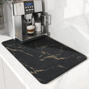 A stainless steel espresso machine with a finished cappuccino sits on a counter alongside the Lovely Home Icon Coffee Machine Mat—an elegant black water-absorbent dish drying pad featuring gold marble-like streaks—set against a white marble backsplash.