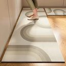 A person stands on the Lovely Home Icon New 1 pc Japanese style soft kitchen diatomite mud floor mat in beige, featuring curved geometric patterns. The super anti-slip and super absorbent runner rug lies on a hardwood floor in a sunlit kitchen, with only the lower legs and feet in suede shoes visible. Another matching modern-style mat from Lovely Home Icon can be seen partially in the background.