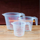 Three Lovely Home Icon plastic measuring cups in 250ml, 500ml, and 1000ml sizes are arranged on a wooden surface against a dark background. Each transparent PVC scale cup features clear red markings for precise measurement, making this 3-piece set of baking liquid measuring beakers essential kitchen tools for cooking and baking tasks.