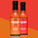 Two bottles of Bravado Spice hot sauce stand side by side. One bears the label "Serrano & Basil Hot Sauce," infusing Italian-inspired flair, while the other showcases Arbol Chili & Garlic. Behind them, bold red and orange diagonal color blocks create a vibrant backdrop—setting the scene for botanical heat that's just right for drizzling on your next pizza.