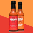 Two bottles of Bravado Spice Co hot sauce—one featuring the Bravado Spice Co Árbol Chili & Garlic Hot Sauce, renowned for its bold Mexican flavors that add zest to any dish, and the other Serrano & Basil—stand side by side on a vibrant red and orange background.