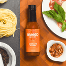 A bottle of Bravado Spice Serrano & Basil Hot Sauce by Bravado Spice rests on a wooden surface, surrounded by pasta, black peppercorns, basil leaves, sliced meat, and a dish of crushed red pepper—the perfect Italian-inspired hot sauce for drizzling over pizza or adding fresh botanical heat to your favorite dishes.