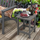The Square Adirondack Side Table 14″ Weather-Resistant HDPE by DirectDeals holds grapes, glasses, and wine beside a matching cushioned chair on a wooden deck surrounded by lush garden plants.