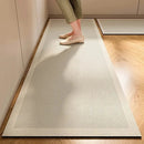 A person stands on the Lovely Home Icon New 1 pc Japanese style soft kitchen diatomite mud floor mat, showcasing only their legs and feet against the wooden flooring. The light-colored, striped rectangular mat combines modern elegance with practicality, offering super anti-slip stability and superior absorbency. In the background near the wall, a smaller matching carpet from Lovely Home Icon adds a cohesive touch to the space.