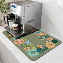 A silver espresso machine sits on the Lovely Home Icon Coffee Machine Mat, a water-absorbent dish drying pad featuring colorful flowers and leaves, adding a modern touch to the white marble countertop. A cup of cappuccino is positioned under the machine’s spout.