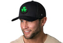 A man in a Haka Hat Shamrock Trucker Hat—black St. Patrick's Day snapback with green shamrock patch—smiles and looks off to the side, sporting short facial hair and a light-colored shirt.