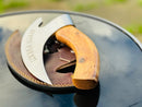 The Texan Knives Vikings Pizza Cutter features a custom handmade 8-inch J2 steel blade with a wooden handle, resting on its brown leather sheath atop a glossy black outdoor surface.