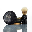 A shaving brush stands next to a ceramic bowl containing The Immaculate Beard Shave Soap Puck, wrapped in black paper. Reflected on a white surface, this set from The Immaculate Beard is an excellent option for those with sensitive skin.